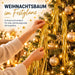 Hände schmücken einen beleuchteten Weihnachtsbaum mit goldenen Girlanden und Kugeln; Werbetext links im Bild.